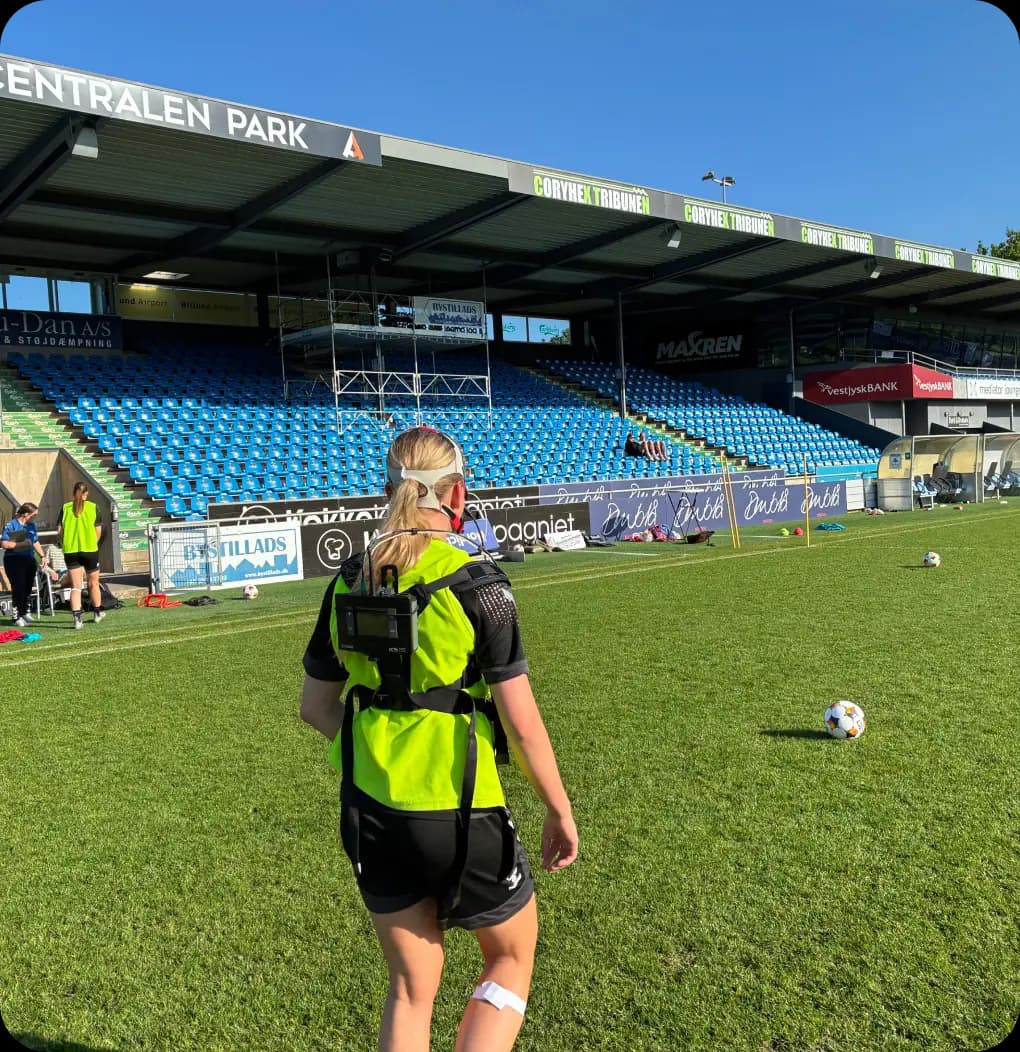 Kvindelig fodboldspiller med GPS-vest under en træning på stadion med blå sæder og solskin.
