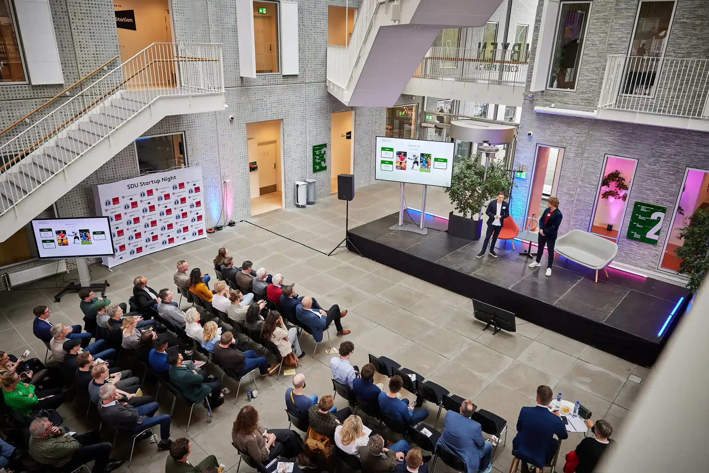 Præsentation under SDU Startup Night i et moderne atrium. To oplægsholdere står på scenen foran et siddende publikum, med store skærme og en bagvæg med sponsorerlogos.